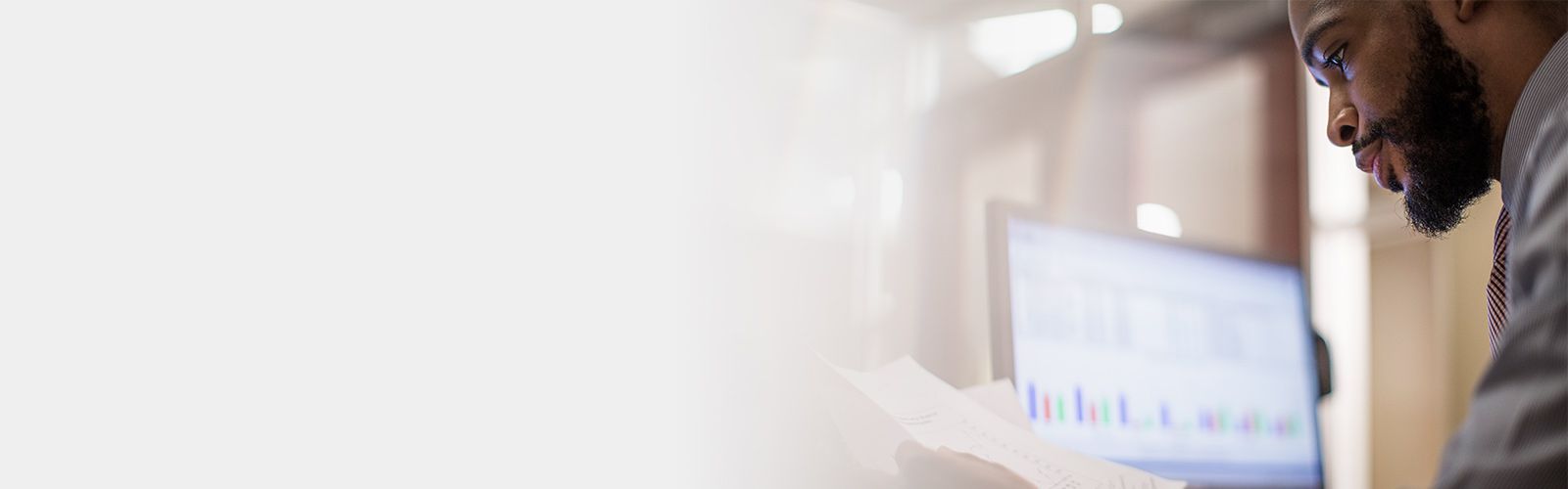 Man looking at paperwork with computer monitor in background.