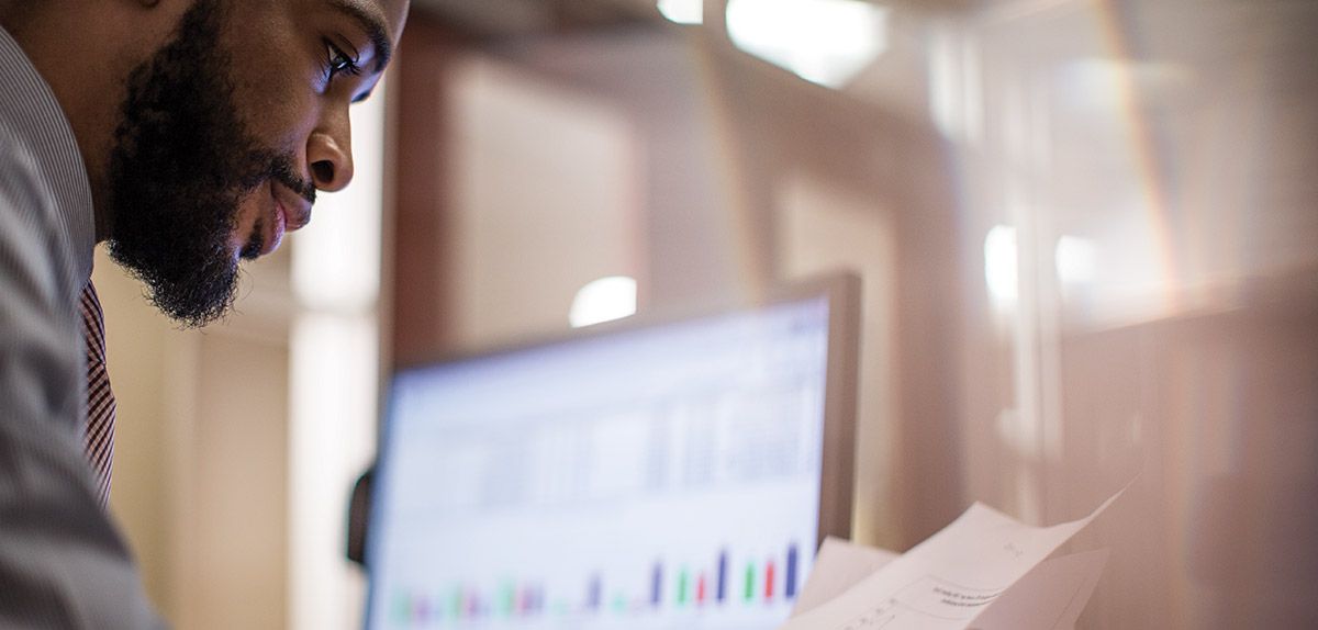 man studying printed documents while sitting next to computer displaying a chart and data.