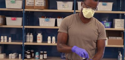 A pharmacist wearing exam gloves and a mask holding a pill bottle in a pharmacy.